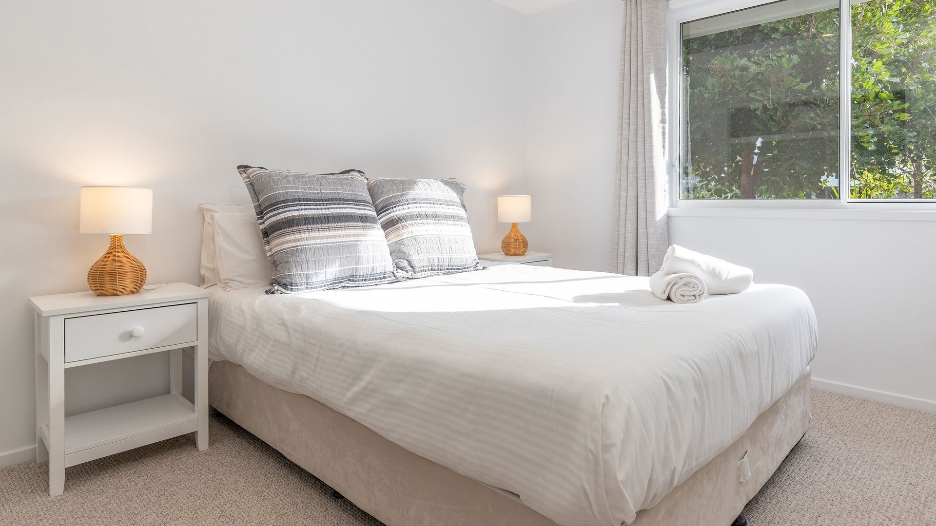 A neatly made bed with white bedding and striped pillows, two bedside tables with lamps, a window with curtains, and a view of greenery outside.