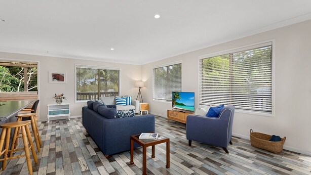 A modern living room with a sofa, armchairs, a coffee table, and a TV. There are large windows with blinds, a dining area with a table and chairs, and a lamp. - Marine Villa @ Smiths Lake (Tarbuck Bay)