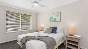 A bedroom with a bed, two bedside tables, a ceiling fan, a window with blinds, and a wall-mounted artwork.