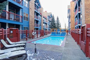 A swimming pool surrounded by multi-story buildings with balconies, a red fence, and lounge chairs with snow on them. - Ski-In Condo, Walk to Gondola | Hot Tubs (Breckenridge)