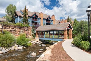 A stone bridge over a stream, leading to a building with a blue facade and a covered walkway. - Ski-In Condo, Walk to Gondola | Hot Tubs (Breckenridge)