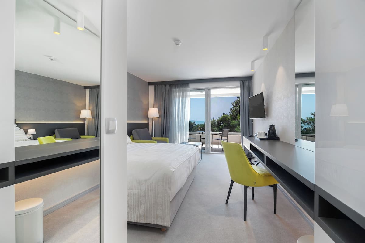 A modern hotel room with a bed, a desk, a chair, a TV, and a balcony with a view.