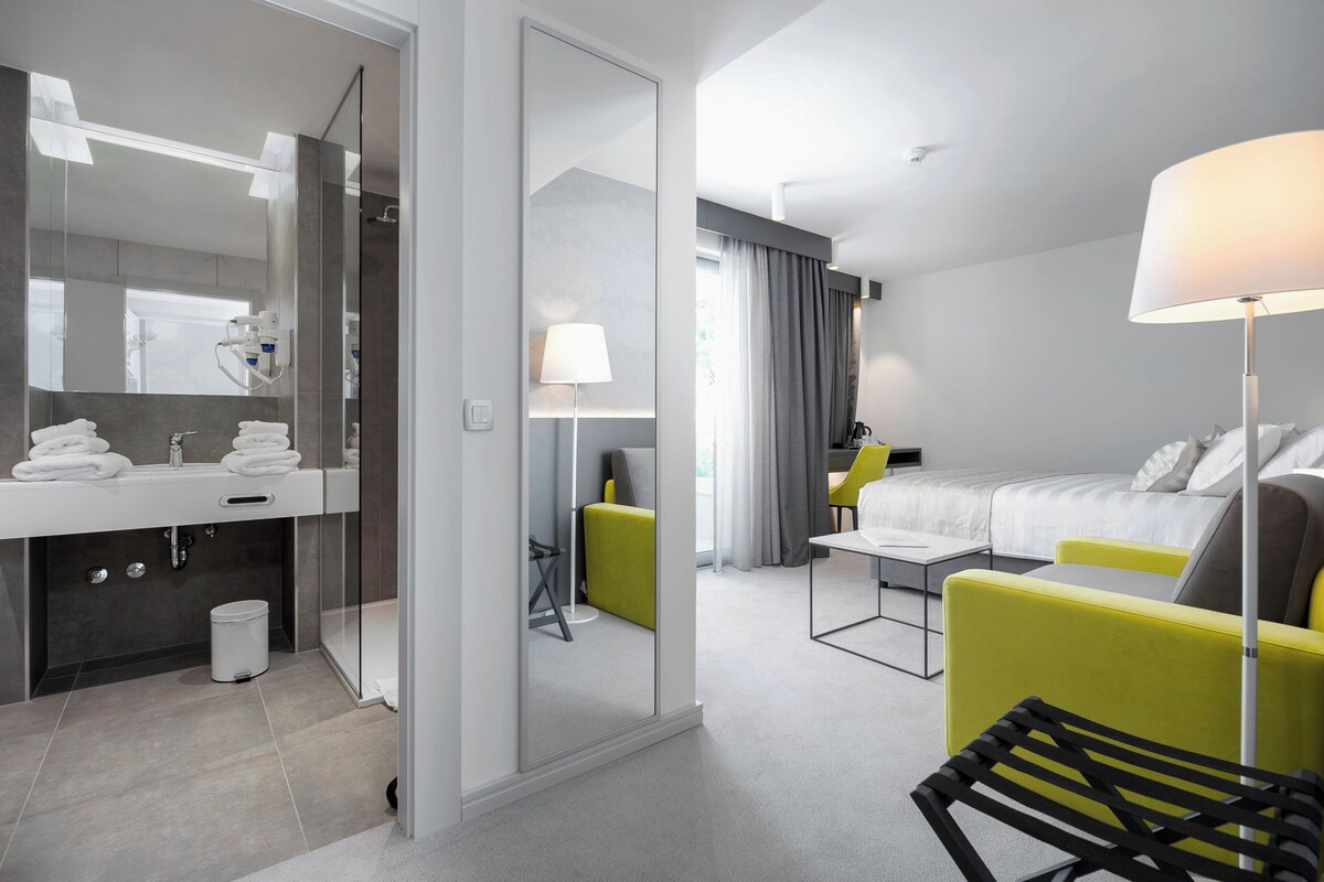 A modern hotel room with a bathroom, a bed, a desk, and a chair.
