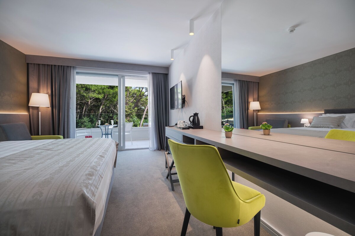 A modern hotel room with a bed, desk, and chairs, a large window with curtains, and a view of greenery outside.