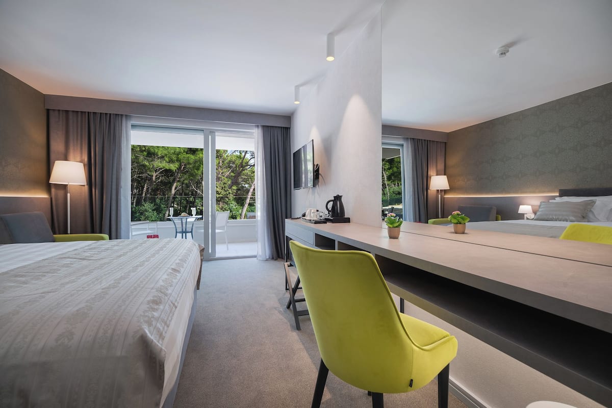 A modern hotel room with a bed, desk, and chairs, a large window with curtains, and a view of greenery outside.
