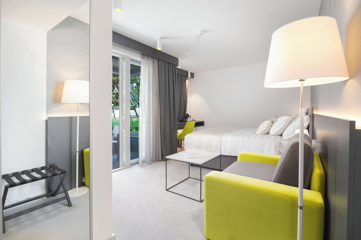Una habitación de hotel moderna con una cama, un sillón amarillo, una mesita y un banco.
