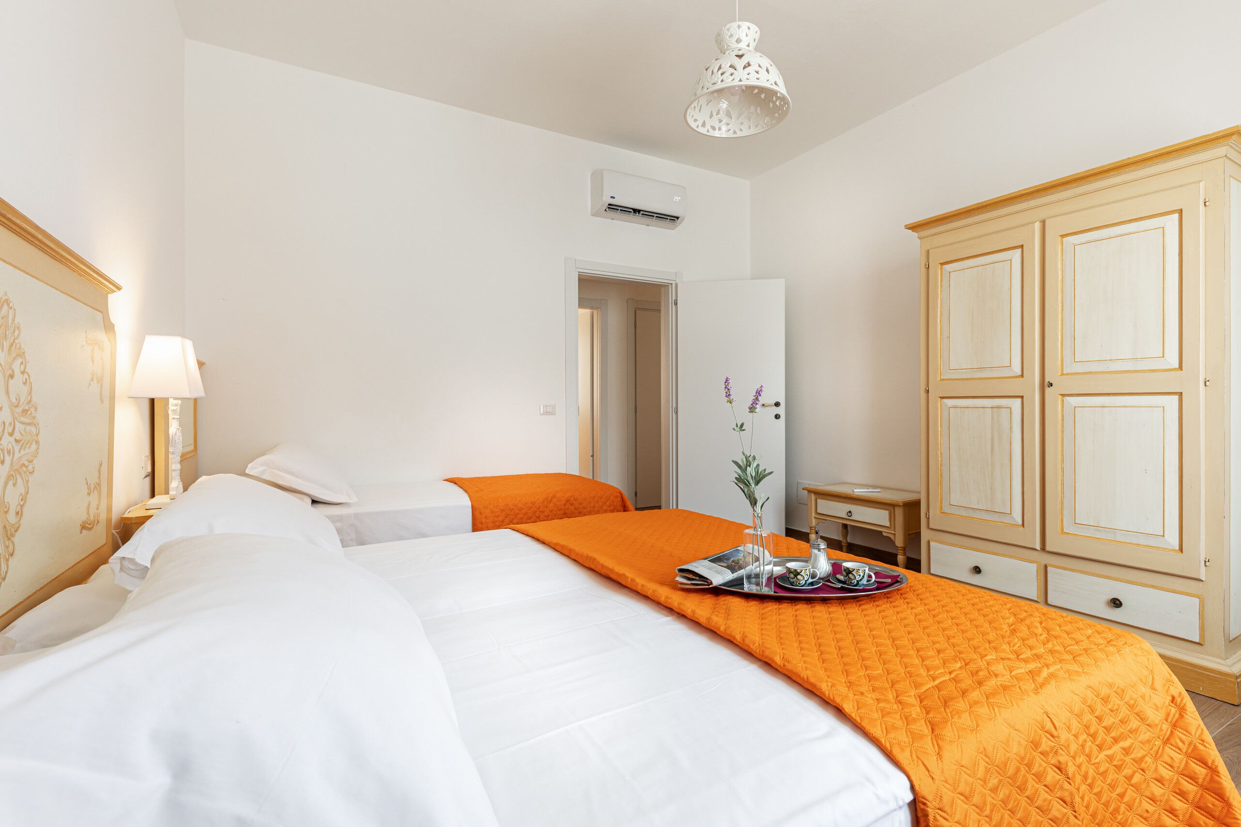 Habitación de hotel con dos camas, una colcha naranja, un armario de madera, una mesita de noche con lámpara y un accesorio de iluminación en el techo.