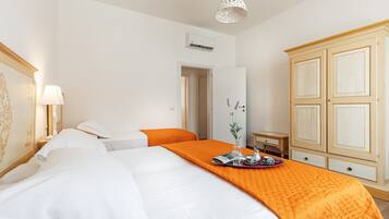 Habitación de hotel con dos camas, una colcha naranja, un armario de madera, una mesita de noche con lámpara y un accesorio de iluminación en el techo.