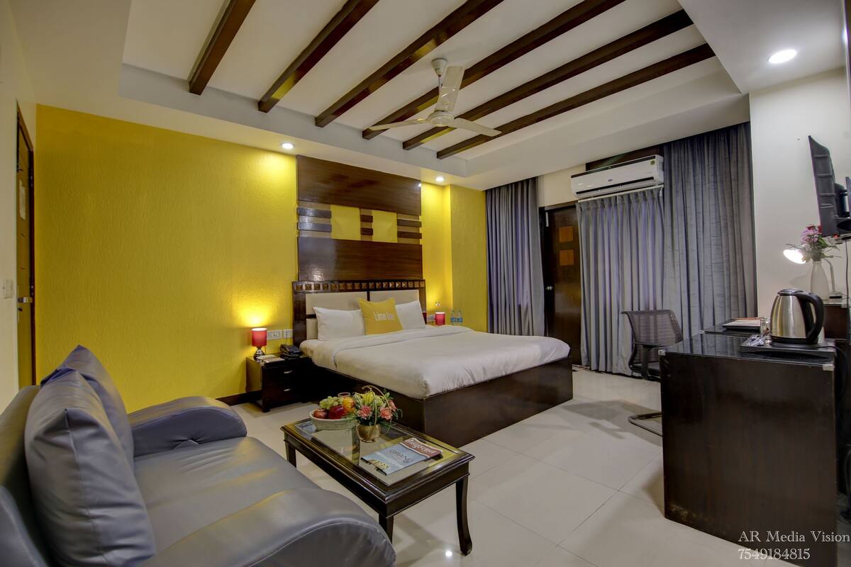 A hotel room with a bed, a sofa, a coffee table with fruits and flowers, a desk, an air conditioner, and a ceiling fan.