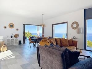 A modern living room with a large sofa, dining area, and large windows offering a view of the sea.