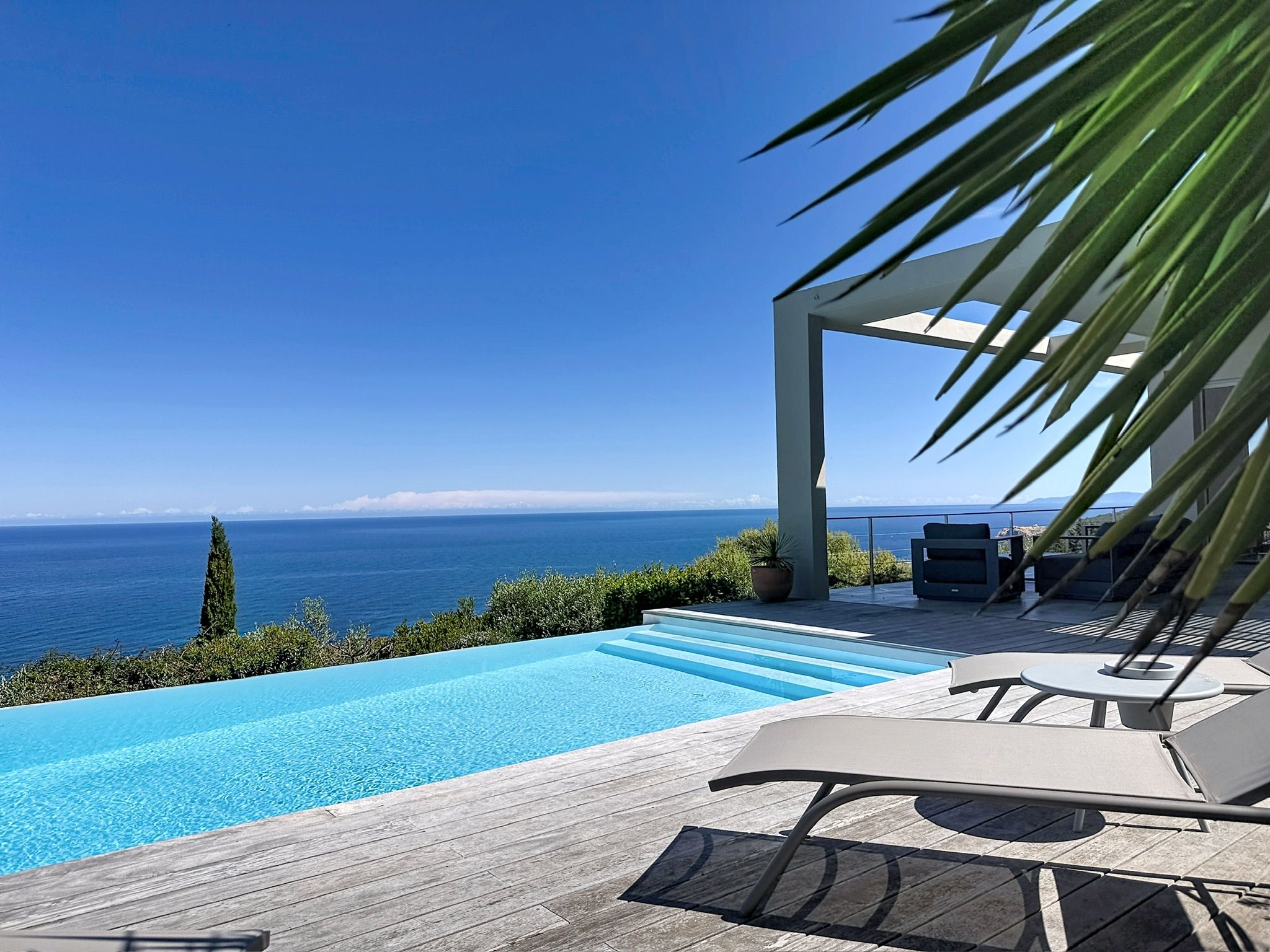 A swimming pool with a view of the sea, a wooden deck, and outdoor furniture.
