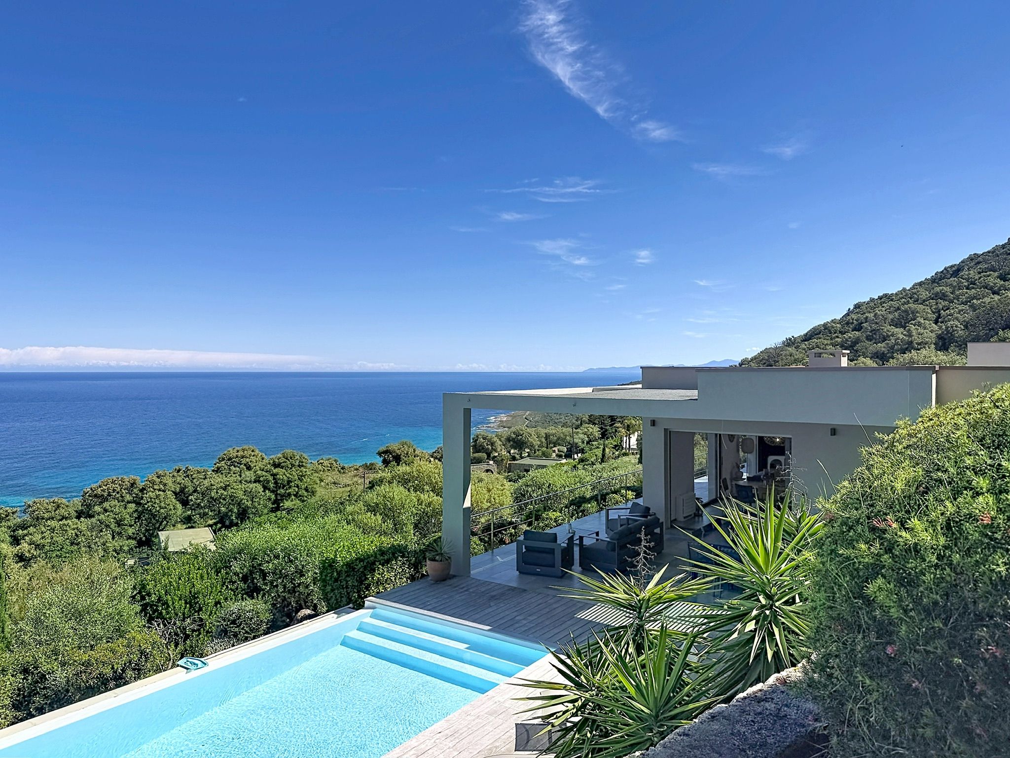 A modern pool area with a view of the sea, surrounded by lush greenery and a hillside.