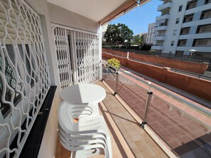 Un balcon avec une table et des chaises en plastique blanc, une rambarde en métal et une vue sur un paysage urbain avec des bâtiments et un ciel dégagé.