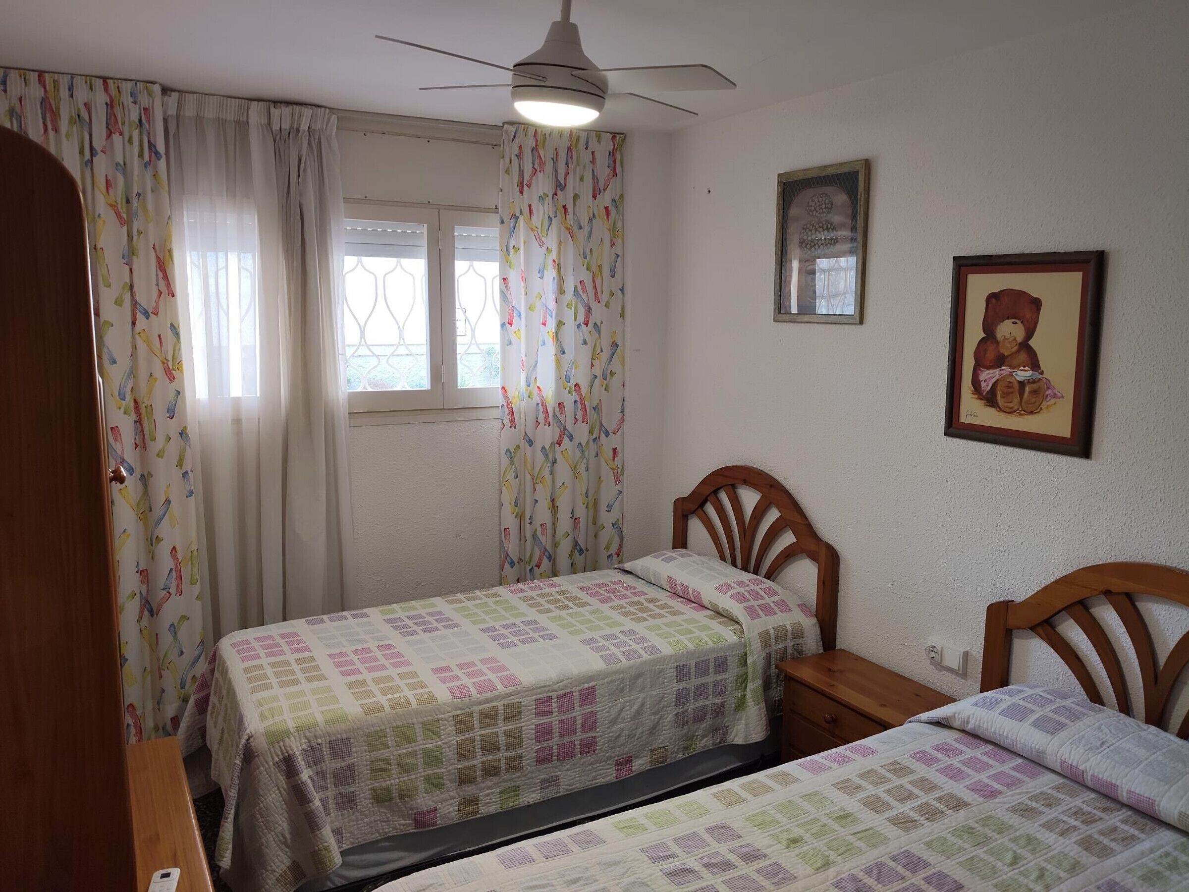 A bedroom with two single beds, a ceiling fan, a window with curtains, and a framed picture on the wall.