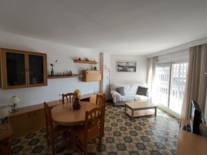 Un salon comprenant une table à manger, des chaises, un canapé, une table basse et un téléviseur. On y trouve également des étagères ornées d’objets décoratifs et une fenêtre avec des rideaux.