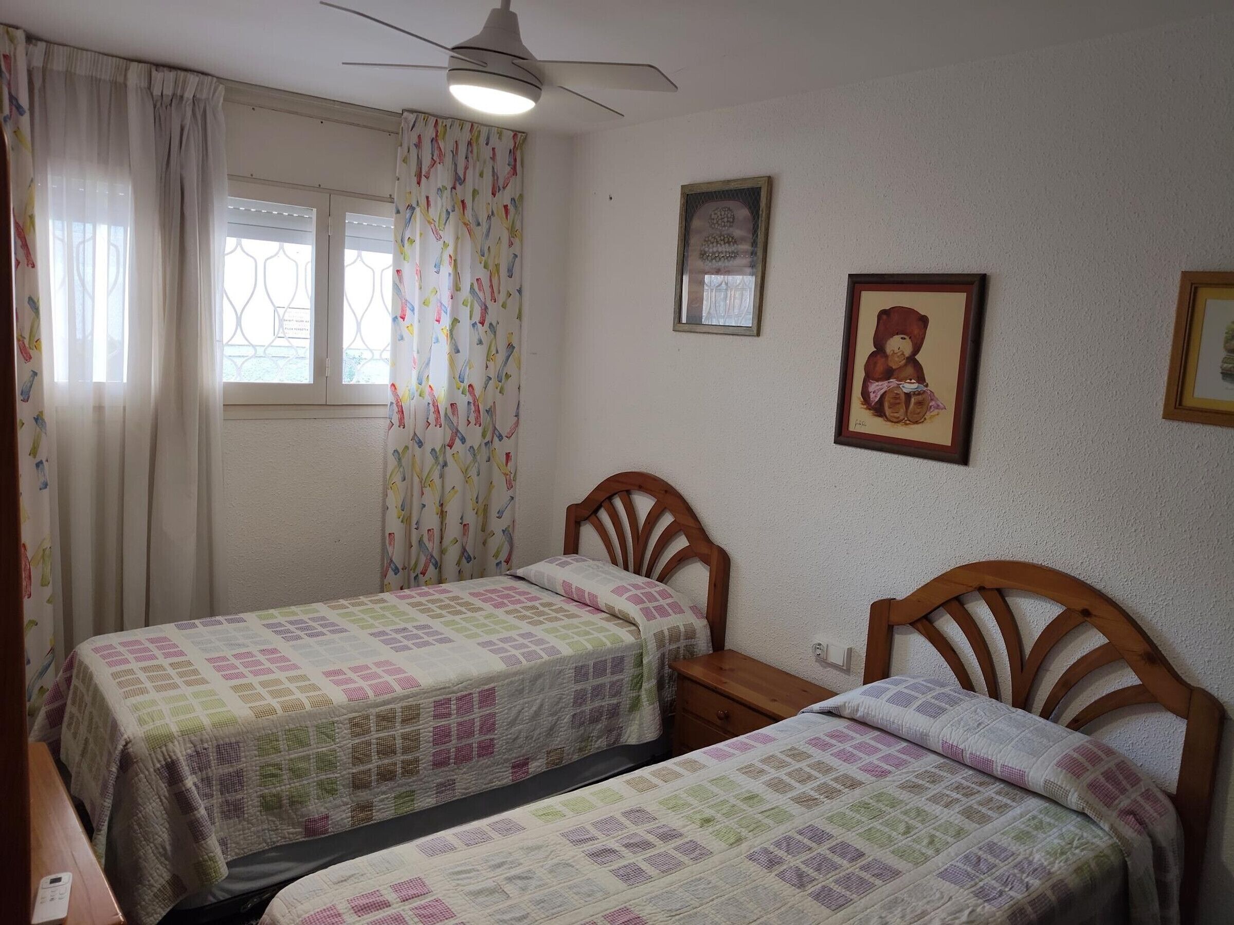 Un dormitorio con dos camas individuales, un ventilador de techo, una ventana con cortinas y un cuadro en la pared.
