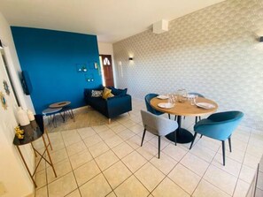 Uma área de refeições com uma mesa redonda e cadeiras, um sofá azul e uma pequena mesa lateral com uma luminária.