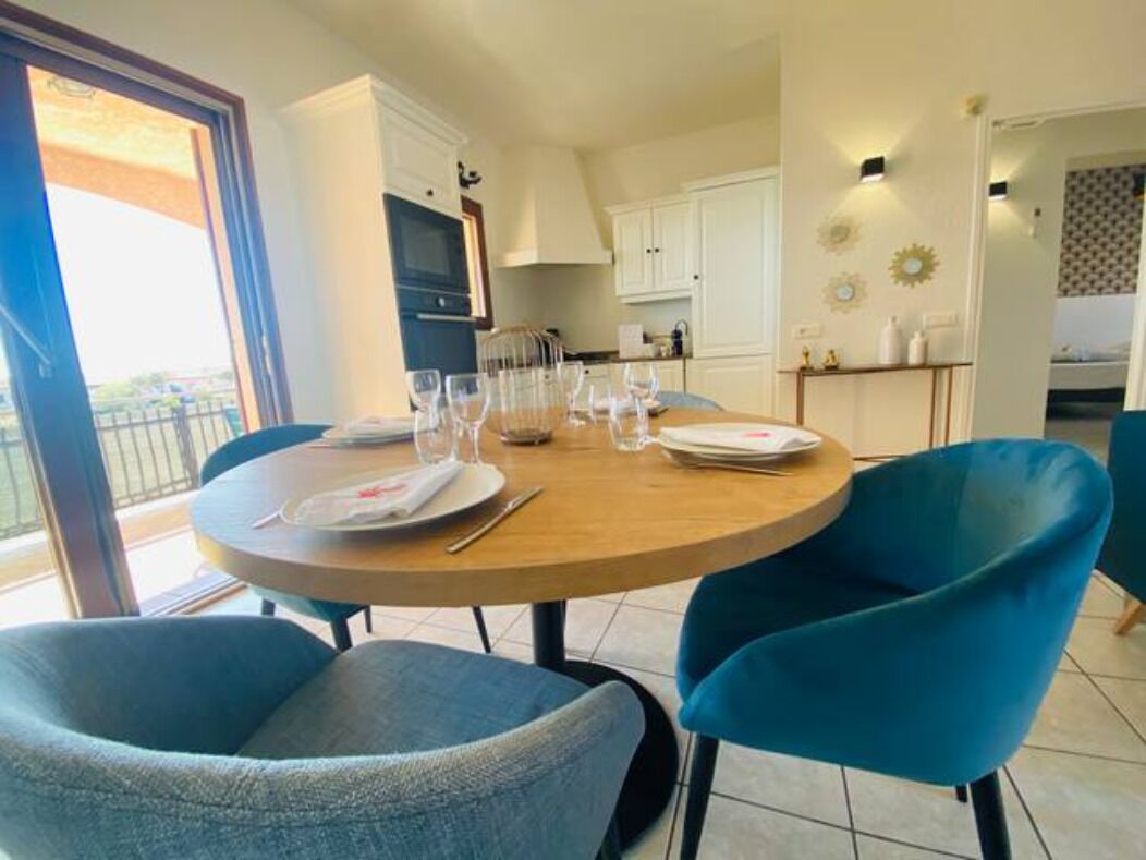 A dining area with a round table set for four, two blue chairs, and a view of the kitchen and balcony in the background.