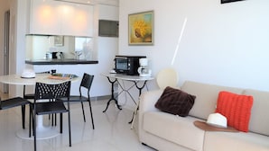 Ein modernes Wohnzimmer mit einer Couch, zwei Sesseln, einem Couchtisch und einer Küchenzeile mit Mikrowelle und Kaffeemaschine.