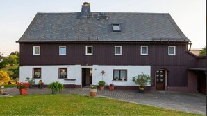Een tweelaags huis met een donkerbruine gevel, een pannendak en een oprit van baksteen. Voor het huis staan verschillende bloempotten.