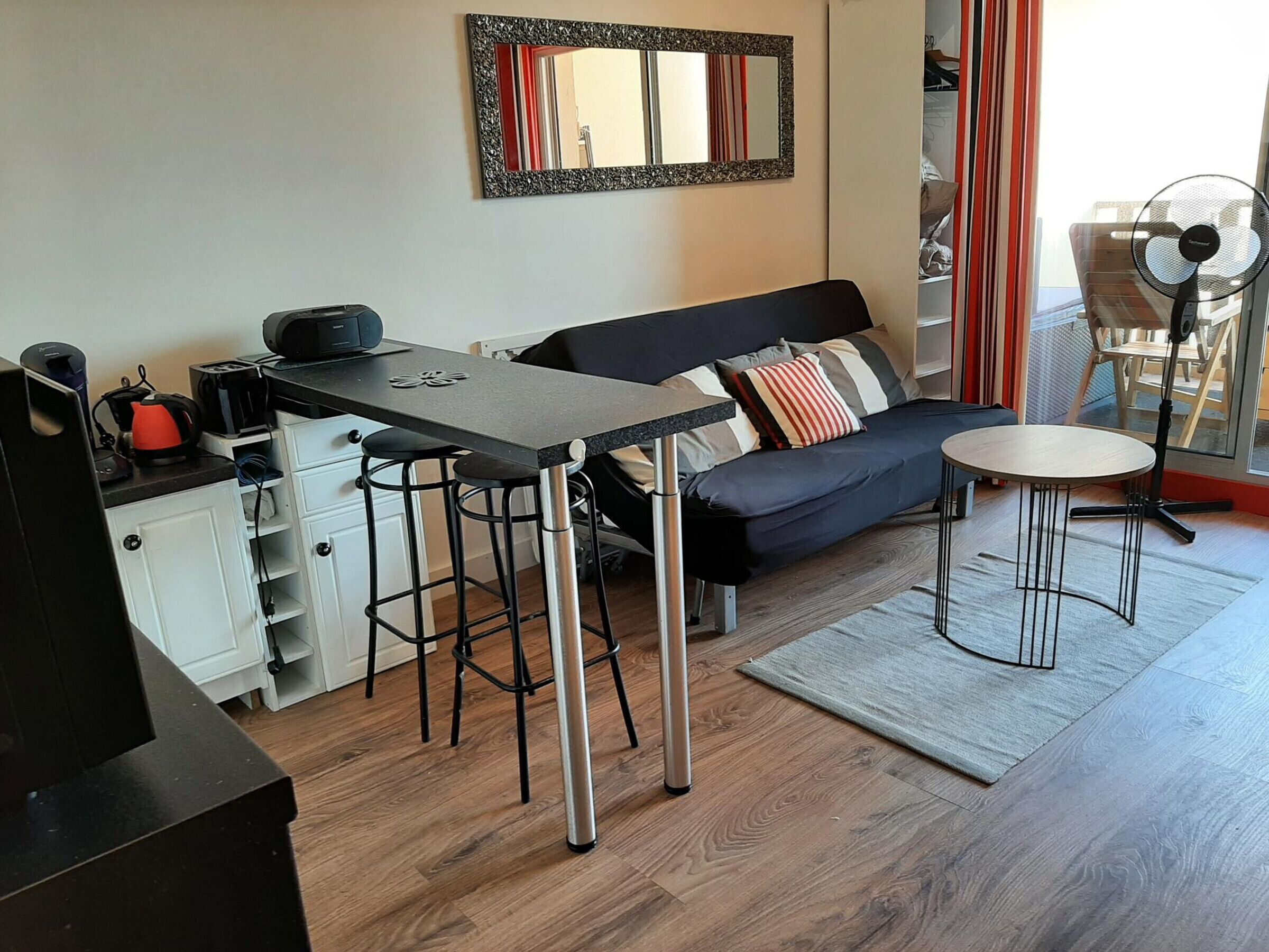 Un espace de vie compact comprenant un coin cuisine, un canapé, une petite table et un ventilateur.
