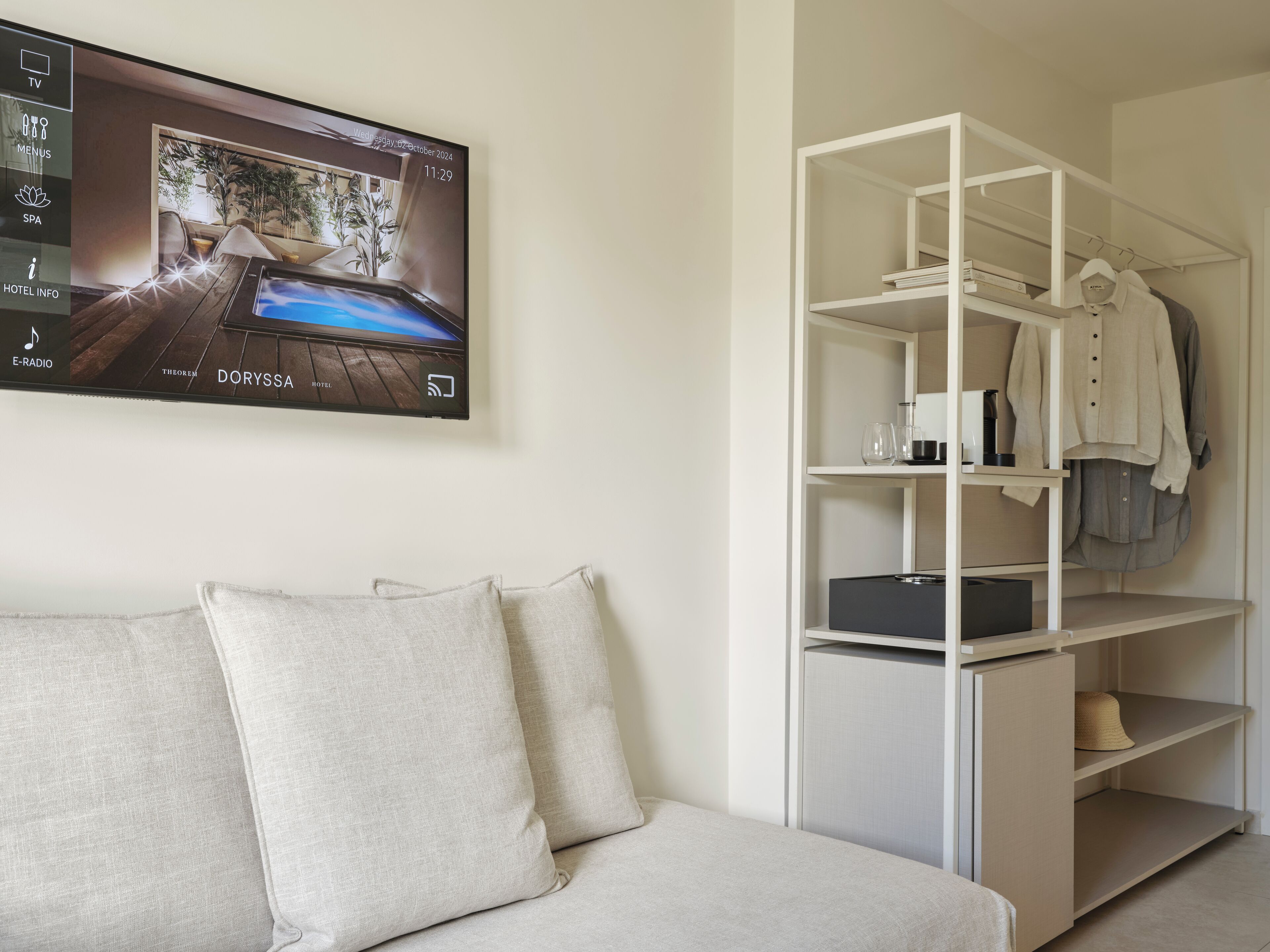 Habitación de hotel moderna con un televisor de pantalla plana mostrando una vista panorámica, un sofá blanco con cojines y un estante blanco con ropa y un sombrero.