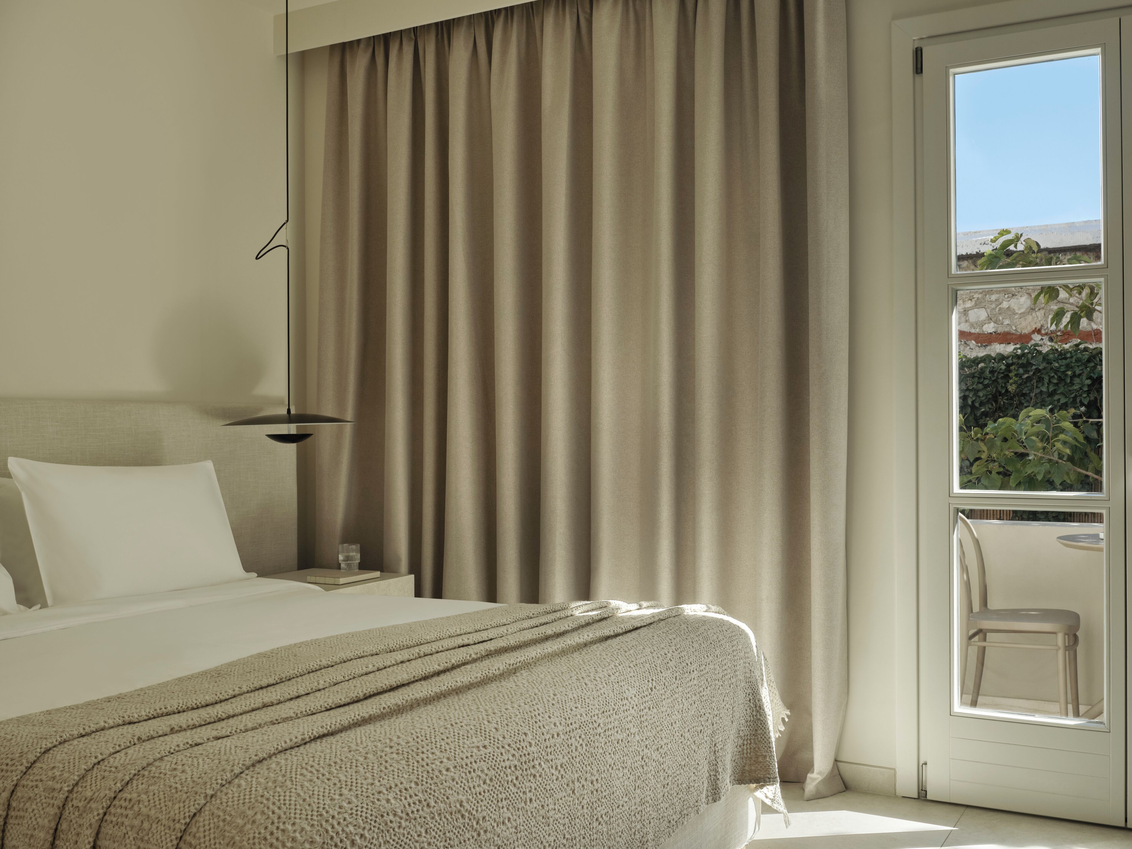 A hotel room with a bed, a nightstand, a lamp, a chair, and a view of greenery through a window.