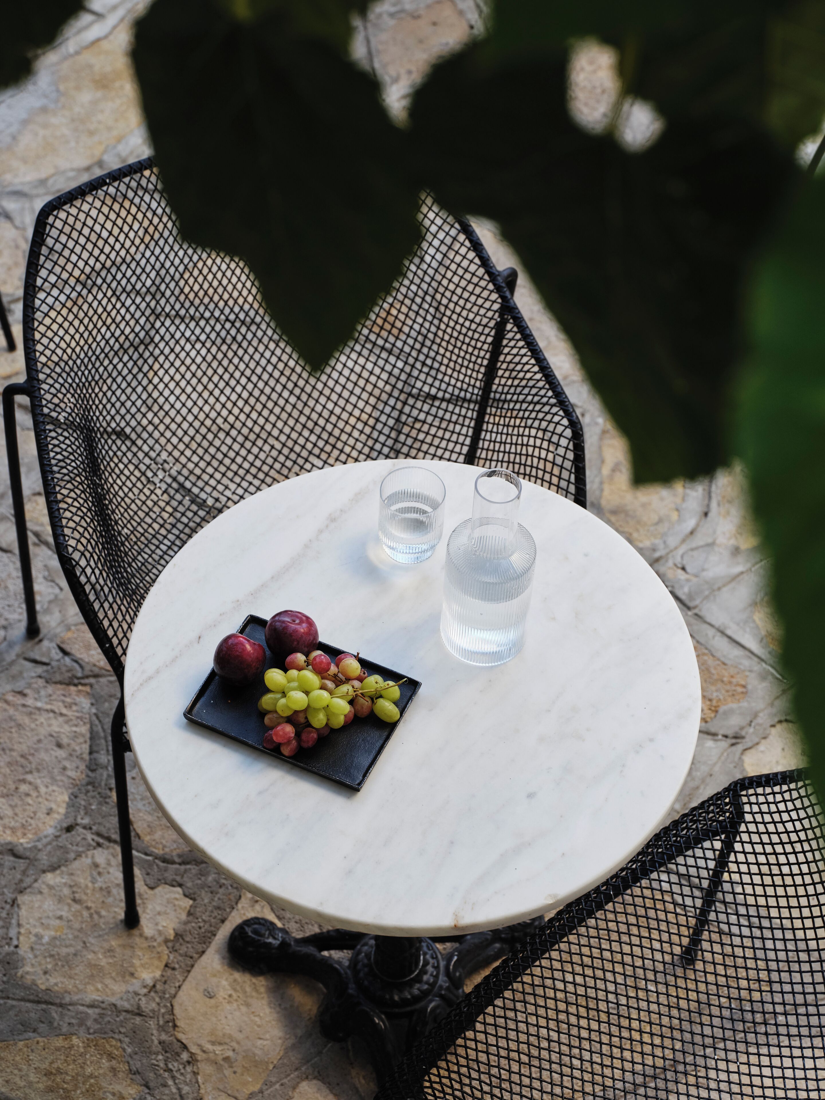 Una mesa redonda de mármol con una silla de metal negro, una bandeja negra con uvas y manzanas, y dos vasos transparentes.