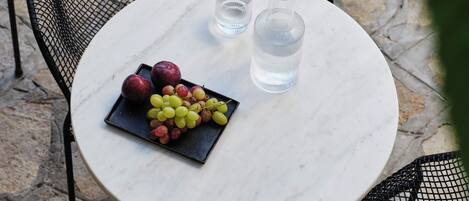 A round marble table with a black metal chair, a black tray with grapes and apples, and two clear glasses.