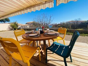 Un'area pranzo all'aperto con un tavolo rotondo in legno, sedie gialle e verdi, e un portico coperto.