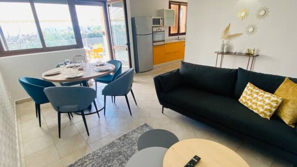 En stue med en sort sofa, et spisebord med stole, og et køkken i baggrunden.