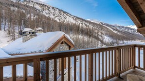 A wooden balcony with a stone building in the background, surrounded by snow-covered mountains and trees. - Luxury chalet, jacuzzi and sauna, sleeps 14. (Val-d'Isère)
