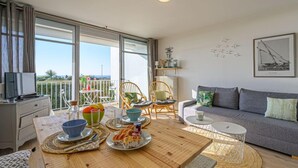 Un soggiorno moderno con un balcone, un divano grigio, un tavolo da pranzo in legno con piatti e ciotole, e un televisore su un comò.