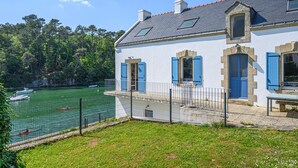 Une maison dotée d’un balcon donnant sur un plan d’eau, d’un jardin clôturé et d’une zone boisée en arrière-plan.