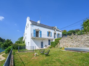 A two-story white house with blue shutters, a stone staircase, and a grassy yard with a chair and a small table. - Glann Aven by Interhome (Le Bono)