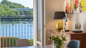 Une pièce avec vue sur un pont et sur l’eau, une table ornée d’un vase de fleurs et une lampe posée sur une commode.