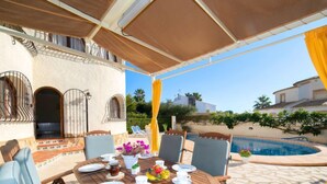 Un área de comedor al aire libre con piscina, suelo de piedra y un porche cubierto.