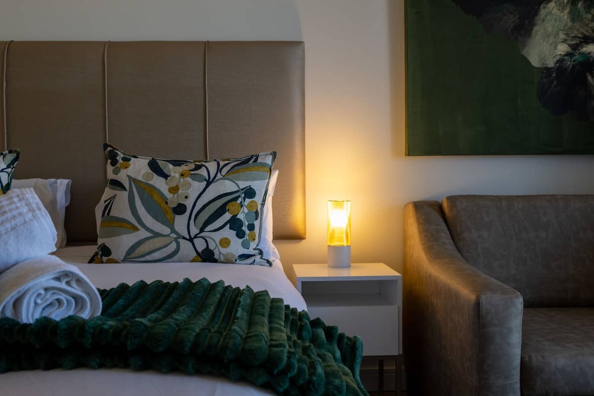 Ein ordentlich bezogenes Bett mit grüner Decke, einem blumigen Kopfkissen und einem aufgerollten weißen Handtuch. Es gibt einen Nachttisch mit einer Lampe, einen braunen Ledersessel und ein Bild an der Wand.