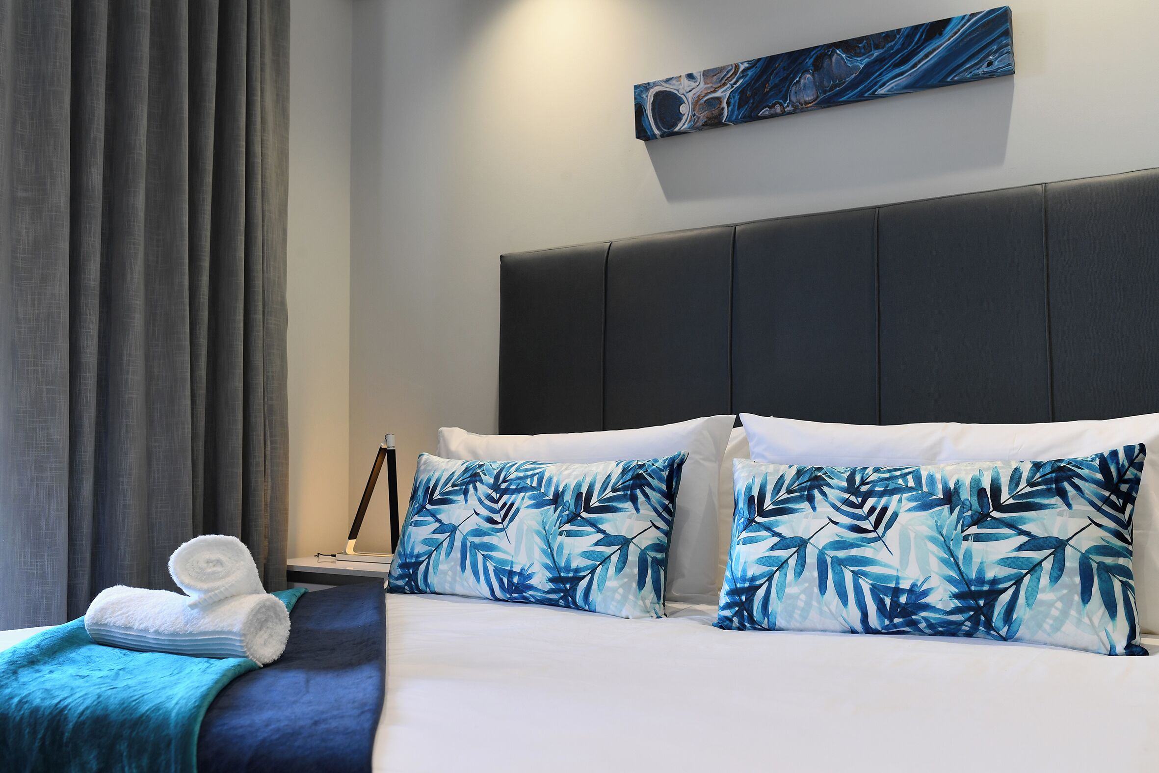 Una camera d'albergo con un letto, cuscini, una coperta, un quadro e un comodino.