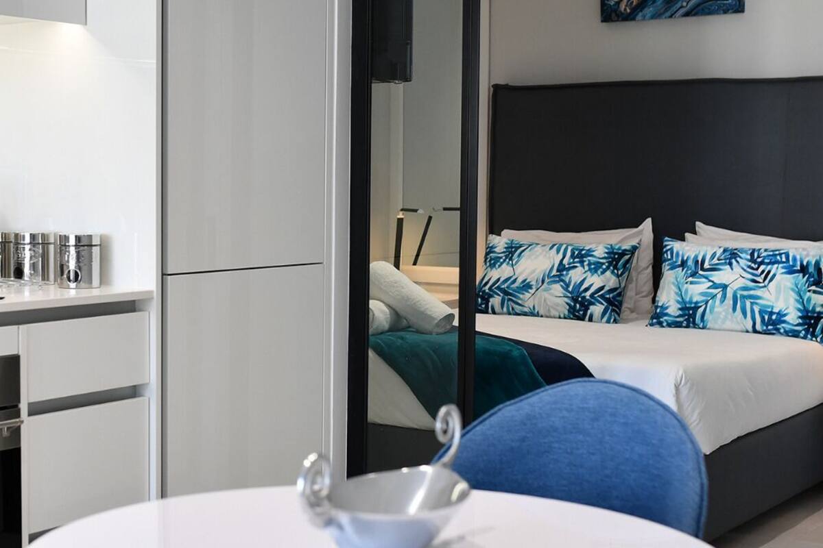 Ein modernes Hotelzimmer mit einer Küchenzeile, einem Bett mit blau-weißen Kissen und einem großen Spiegel, der den Raum reflektiert.