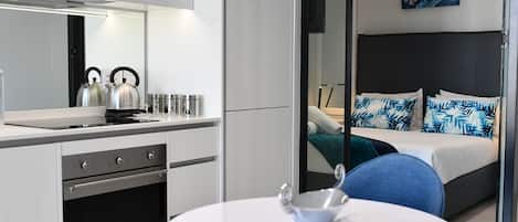 Una habitación de hotel moderna con kitchenette, una cama con almohadas estampadas en azul y blanco, y un espejo grande que refleja la habitación.