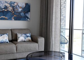 Un salon moderne avec un canapé gris, deux coussins décoratifs, une grande peinture abstraite et une table en verre.