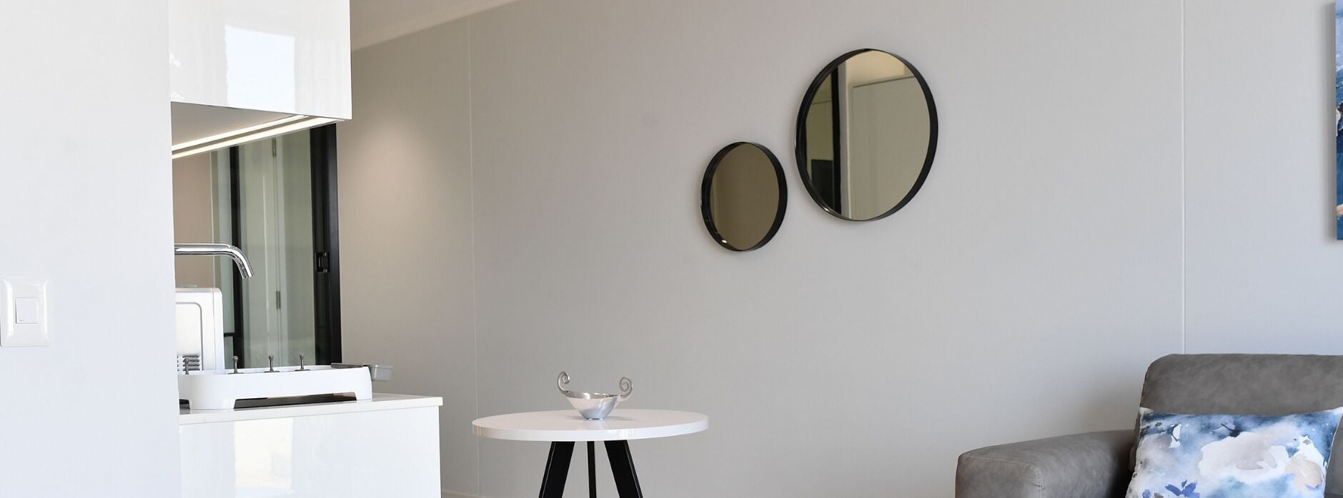 Una habitación moderna y minimalista con un tocador blanco, dos espejos montados en la pared, una pequeña mesa redonda con un cuenco decorativo y un sofá gris con un cojín azul.