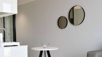 Een moderne, minimalistische kamer met een witte wastafel, twee spiegels aan de muur, een klein rond tafeltje met een decoratieve schaal, en een grijze bank met een blauw kussen.
