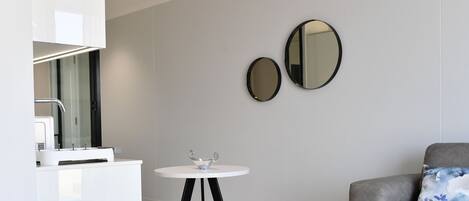 Una habitación moderna y minimalista con un tocador blanco, dos espejos montados en la pared, una pequeña mesa redonda con un cuenco decorativo y un sofá gris con un cojín azul.
