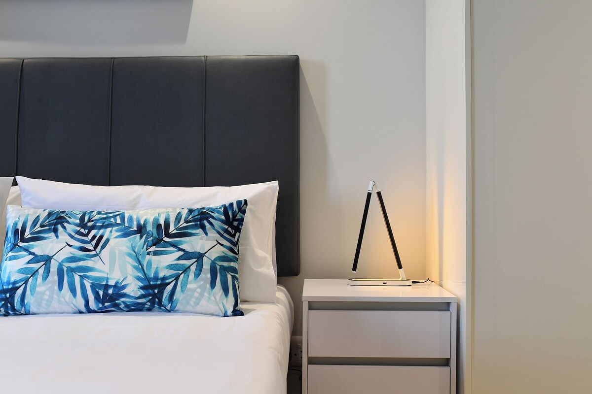 Een bed met een donker hoofdbord, wit beddengoed en een kussen met een bladmotief. Er staat een nachtkastje met een lamp en een klein elektronisch apparaat.