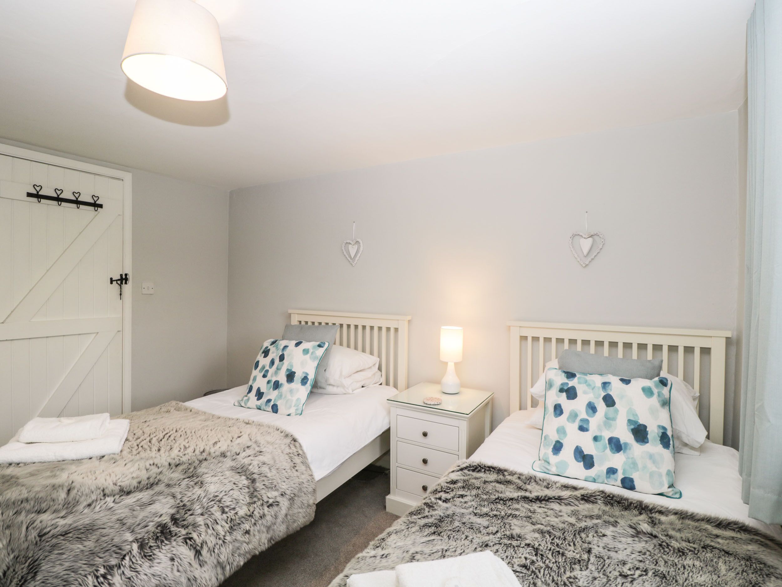 Een slaapkamer met twee bedden, elk voorzien van een wit hoofdeinde en een nachtkastje met een lamp. Er hangen hartvormige decoraties aan de muur en er is een witte deur met een klink.