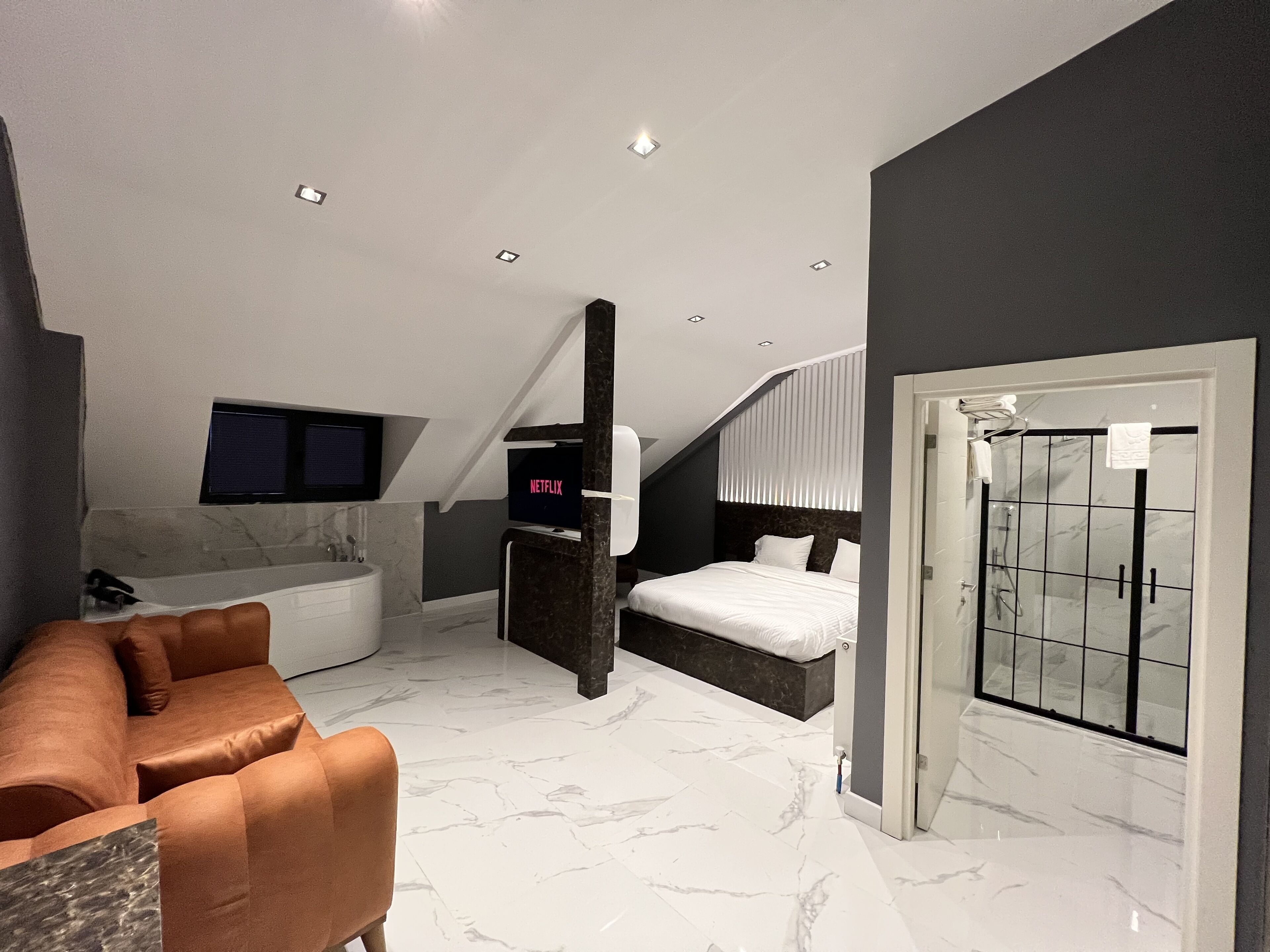 Un dormitorio moderno con una cama grande, televisor integrado, baño con ducha de vidrio y un rincón de descanso con sofá.