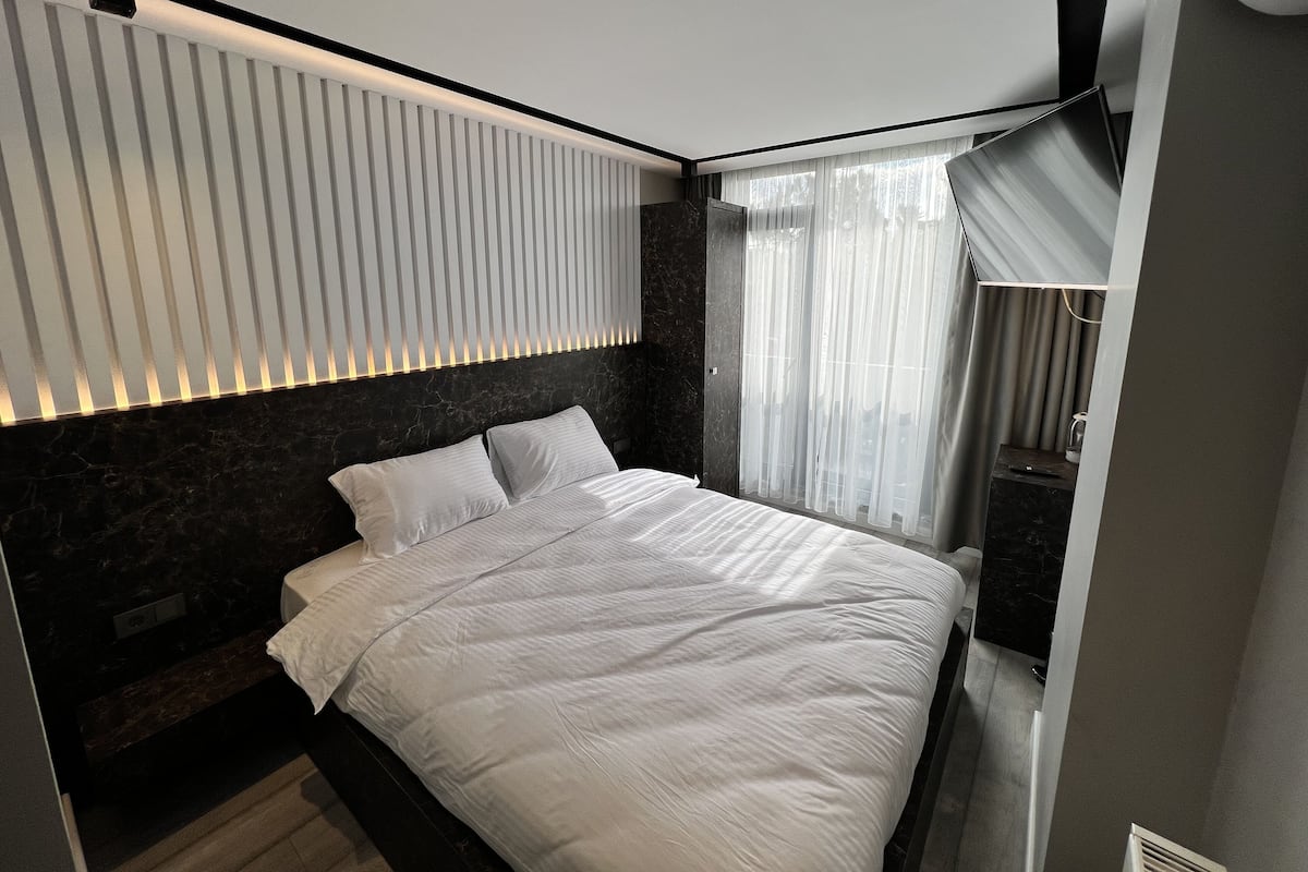 A modern bedroom with a bed, a headboard with vertical slats, a bedside table, and a window with sheer curtains.