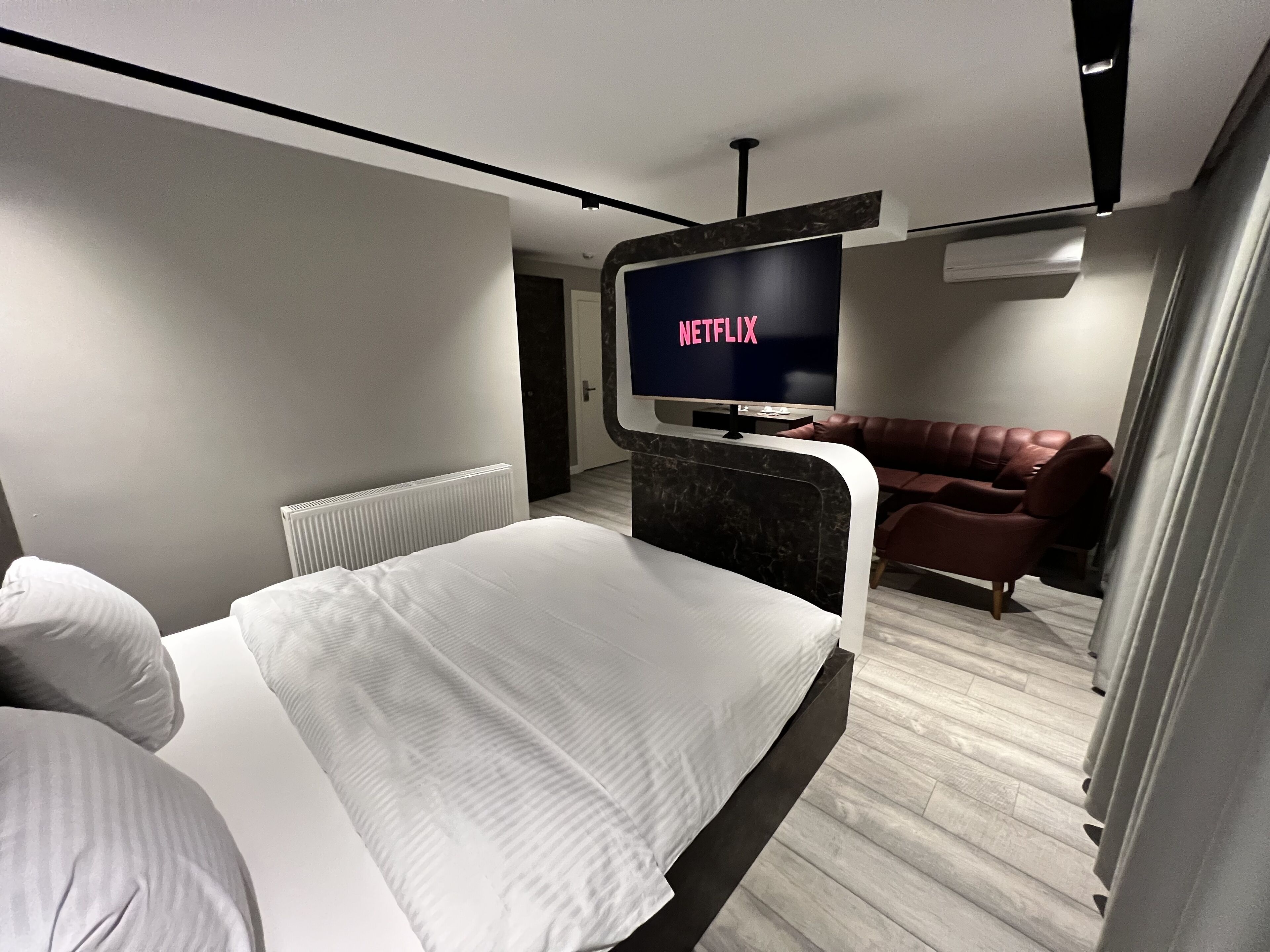 A modern hotel room with a large bed, a TV displaying 'NETFLIX', a leather sofa, and a radiator.